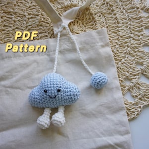 Puede incluir: Una nube de ganchillo azul claro con una cara sonriente y piernas blancas, unida a una cuerda blanca y una bola azul claro. La nube cuelga de un bolso tote beige. El texto "PDF Pattern" es visible.