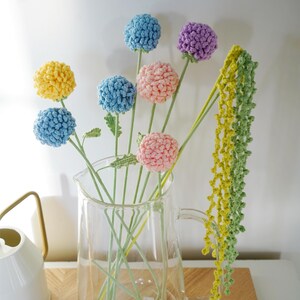 May include: A selection of handmade crochet flowers in a clear glass jug. The flowers are in various colours, including yellow, blue, pink, and purple. The stems are green, and the flowers have a textured, pom-pom-like appearance. A decorative piece.