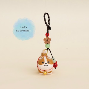 May include: A small, brown and white ceramic cat charm with a gold bell, hanging from a black cord with decorative beads and a wooden flower. The charm is suspended against a light background, with the text "LAZY ELEPHANT" in a blue circle.