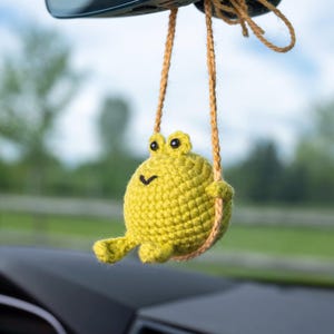 May include: A handmade, yellow crochet frog car charm hanging from a tan string. The frog has black eyes and a small, smiling mouth. The background is blurred, showing a green field and a blue sky.