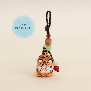 Könnte beinhalten: Eine kleine, orangefarbene Tigerfigur mit schwarzen Streifen und einem lächelnden Gesicht. Der Tiger ist an einer schwarzen Schnur mit dekorativen Perlen befestigt. Der Text "LAZY ELEPHANT" befindet sich in einem blauen Aquarellkreis im Hintergrund.