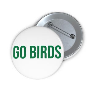 Puede incluir: Dos insignias redondas blancas con el texto verde "GO BIRDS". Una de las insignias está boca arriba, la otra muestra la parte posterior del alfiler. Estas insignias son probablemente para un aficionado al deporte.