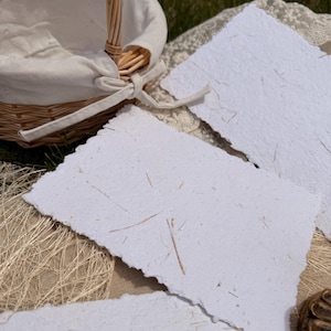 May include: A wicker basket with a white fabric lining and a tied bow, next to several handmade white paper sheets with rough edges. The paper has visible fibres, suggesting a natural, textured quality. The scene is set outdoors.
