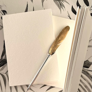 May include: A stationery set featuring a gold feather pen, blank cream-colored cards, and a stack of notepaper. The pen has a silver shaft and a decorative top. The background is a floral pattern in black and white.