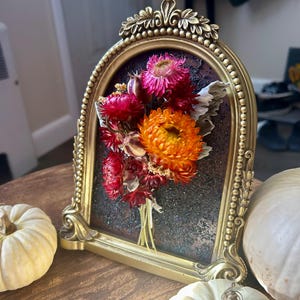 K&ouml;nnte beinhalten: Ein goldfarbener, gerahmter Blumenstrau&szlig; mit getrockneten Blumen in Rot-, Rosa- und Oranget&ouml;nen. Der Rahmen hat ein verziertes Design und ist vor einem dunklen Hintergrund platziert. Zwei wei&szlig;e K&uuml;rbisse befinden sich im Vordergrund.