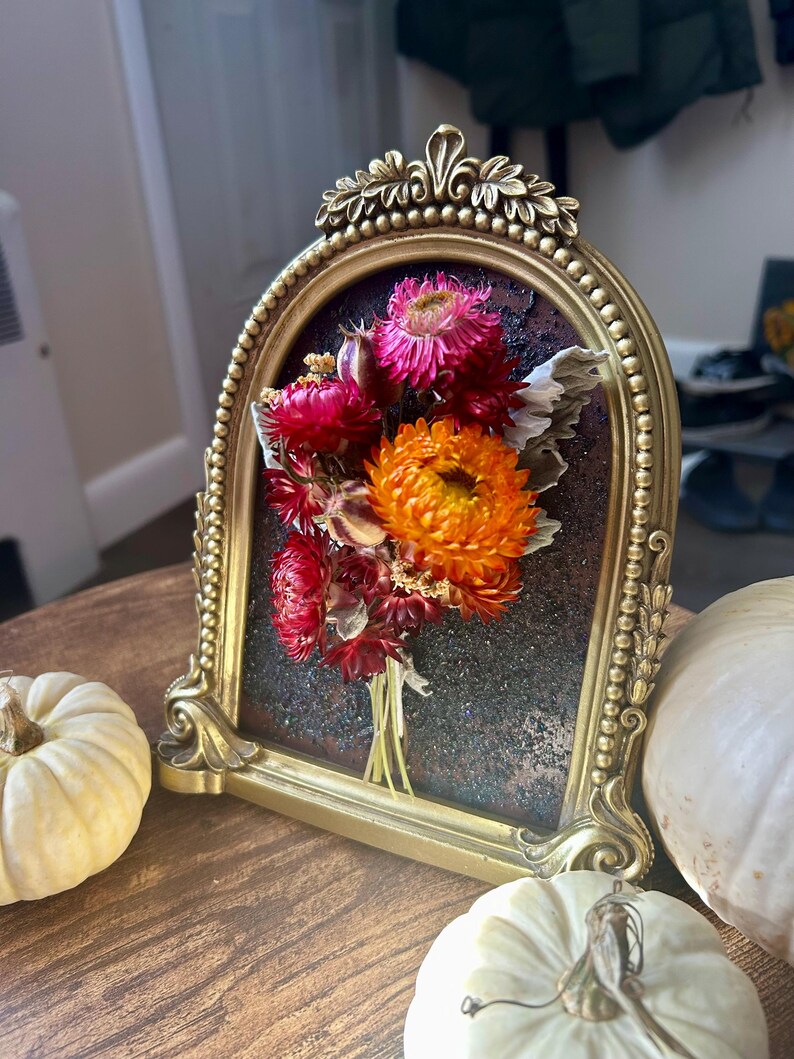 K&ouml;nnte beinhalten: Ein goldfarbener, gerahmter Blumenstrau&szlig; mit getrockneten Blumen in Rot-, Rosa- und Oranget&ouml;nen. Der Rahmen hat ein verziertes Design und ist vor einem dunklen Hintergrund platziert. Zwei wei&szlig;e K&uuml;rbisse befinden sich im Vordergrund.