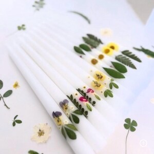 K&ouml;nnte beinhalten: Wei&szlig;e Spitzkerzen, verziert mit gepressten Blumen und Gr&uuml;n. Die Kerzen zeigen eine Vielzahl farbenfroher Blumenarrangements, darunter gelbe, violette und rosa Bl&uuml;ten sowie gr&uuml;ne Bl&auml;tter. Die Kerzen sind auf einer wei&szlig;en Oberfl&auml;che angeordnet.