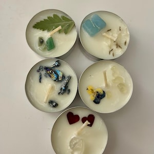May include: Five tealight candles with various natural elements embedded in the wax. The candles feature a green leaf, a blue stone, a iridescent stone, a yellow flower, and two red hearts.
