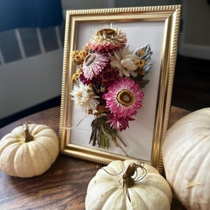 K&ouml;nnte beinhalten: Ein gerahmtes Blumenarrangement mit getrockneten Blumen in Rosa, Wei&szlig; und Gelb. Der goldene Rahmen steht auf einem Holztisch, zusammen mit drei kleinen wei&szlig;en K&uuml;rbissen, f&uuml;r eine herbstliche Dekoration.