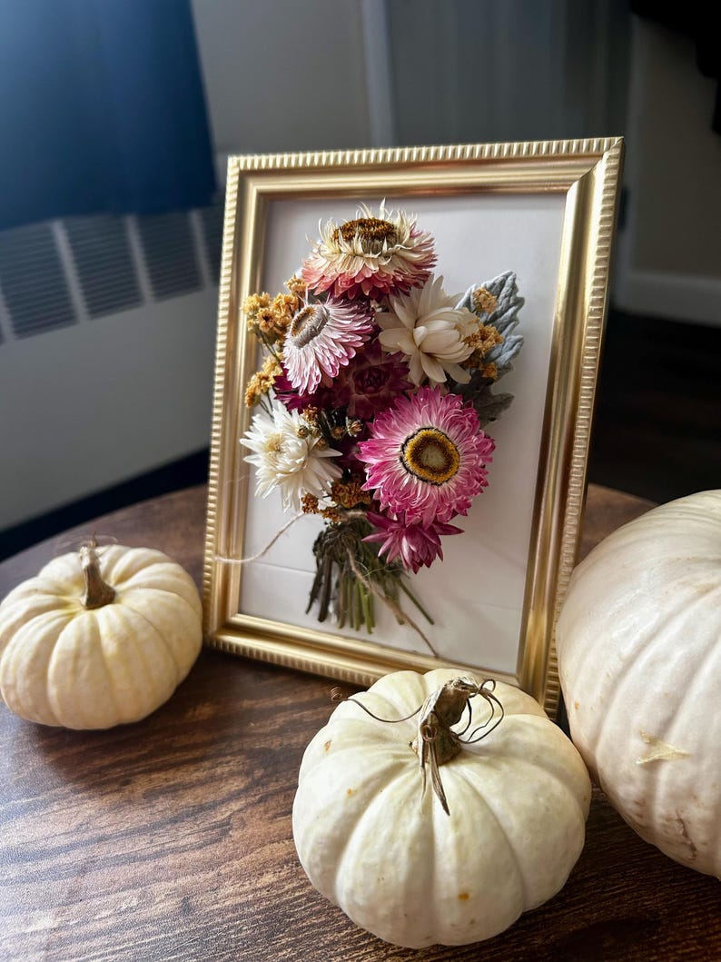 K&ouml;nnte beinhalten: Ein gerahmtes Blumenarrangement mit getrockneten Blumen in Rosa, Wei&szlig; und Gelb. Der goldene Rahmen steht auf einem Holztisch, zusammen mit drei kleinen wei&szlig;en K&uuml;rbissen, f&uuml;r eine herbstliche Dekoration.