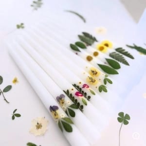 May include: White taper candles decorated with pressed flowers and greenery. The candles feature a variety of floral designs, including yellow, purple, and pink blossoms, and green leaves. The candles are arranged on a white surface.