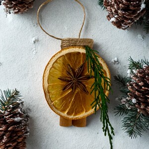 K&ouml;nnte beinhalten: Handgefertigter Weihnachtsschmuck mit einer getrockneten Orangenscheibe, Sternanis und einem Zweig Gr&uuml;n. Der Schmuck ist mit Zimtstangen und Bindfaden verziert, umgeben von Tannenzapfen und Tannenzweigen, was ein festliches Thema ergibt.