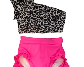 May include: A two-piece swimsuit with a leopard print top and hot pink bottoms. The top has one shoulder strap and short sleeves. The bottoms have a ruffled hem.