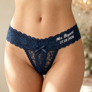 Op de afbeelding: Marineblauwe kanten string met een klein strikje in het midden en witte geborduurde tekst aan de rechterkant met de tekst Mrs. Beyant 07.09.2026. Het kledingstuk is gemaakt van kant met bloemmotief en een geschulpte rand.