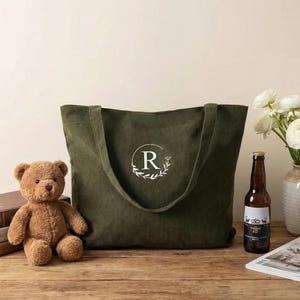 Pode incluir: Uma bolsa tote de veludo cotelê verde-oliva com um design bordado em branco da letra R dentro de uma coroa de louros circular. A bolsa possui duas alças de ombro e uma superfície de tecido texturizado, posicionada entre um urso de pelúcia e uma garrafa.