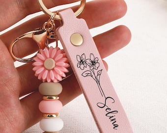 May include: A light pink leather keyring featuring a gold metal ring and clasp, a flower-shaped silicone bead, three round silicone and wooden beads, and a rectangular leather strap printed with a floral line drawing and the name Selina.