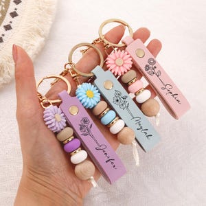 May include: Three personalized keychains featuring a flat leather strap with a gold-tone metal snap, a gold keyring, a silicone flower charm, and three round beads in matching colors. Each strap is printed with a floral line art design and a name.