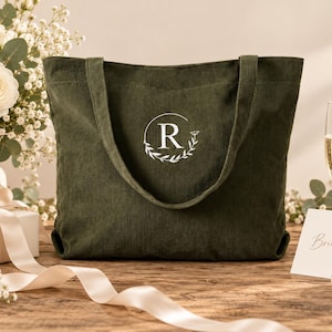 May include: A dark olive green corduroy tote bag featuring a white embroidered design on the front. The design consists of a capital letter R enclosed within a circular frame decorated with delicate leaves and a small flower on the right side.