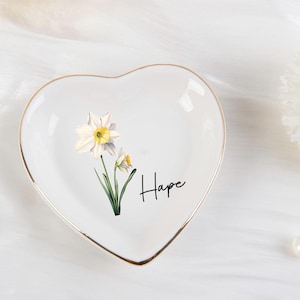 K&ouml;nnte beinhalten: Wei&szlig;es herzf&ouml;rmiges Schmucksch&auml;lchen mit goldenem Rand, mit einem Blumendesign aus Narzissen und dem Wort "Hope" in schwarzer Schrift. Die Schale befindet sich auf einer wei&szlig;en Oberfl&auml;che, mit einer Perlenkette und einer wei&szlig;en Blume im Hintergrund.