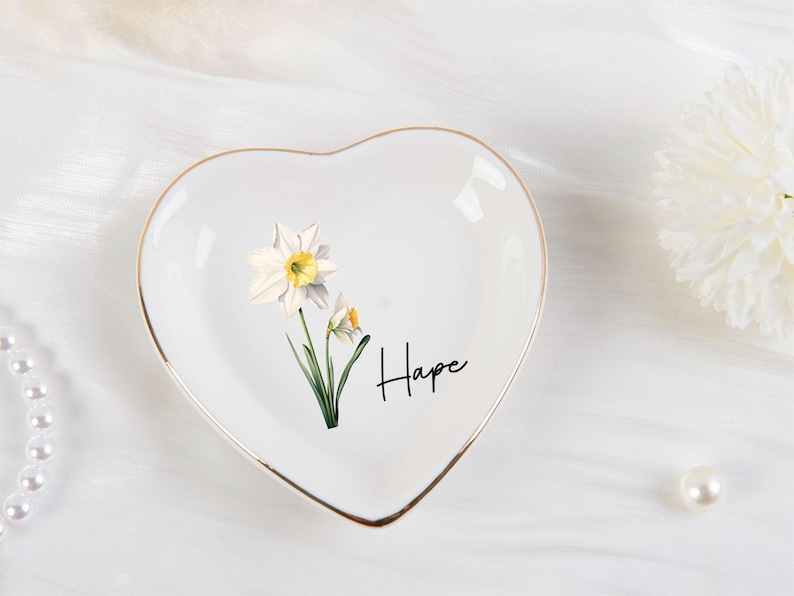 K&ouml;nnte beinhalten: Wei&szlig;es herzf&ouml;rmiges Schmucksch&auml;lchen mit goldenem Rand, mit einem Blumendesign aus Narzissen und dem Wort "Hope" in schwarzer Schrift. Die Schale befindet sich auf einer wei&szlig;en Oberfl&auml;che, mit einer Perlenkette und einer wei&szlig;en Blume im Hintergrund.