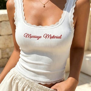 Op de afbeelding: Een wit geribbelde tanktop met een ronde halslijn en armsgaten afgewerkt met delicaat wit kant. In het midden van de borst staat de tekst Marriage Material in een donkerrood cursief lettertype. Het kledingstuk heeft een nauwsluitend model.