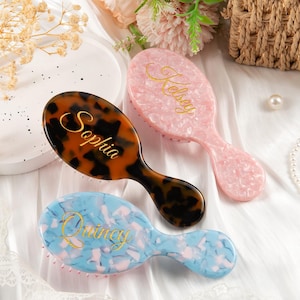May include: Three oval hairbrushes with personalized gold script lettering. One brush features a brown and black tortoiseshell pattern with the name Sophia, one is light pink with the name Kelsey, and one is light blue with the name Quincy.