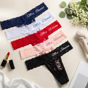 K&ouml;nnte beinhalten: F&uuml;nf Spitzen-Tangas in Marineblau, Wei&szlig;, Rot, Pfirsich und Schwarz, jeweils mit einer kleinen Schleife und wei&szlig;em Sticktext: Mrs. Beyant, Mrs. Daniel, Mrs. Wilson, Mrs. Harper und Mrs. Johnson. Die Kleidungsst&uuml;cke liegen diagonal.
