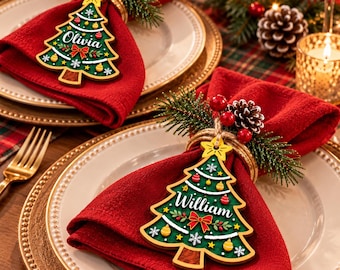 May include: Two red cloth napkins folded on white plates, each secured with a wooden napkin ring featuring a green Christmas tree ornament. The ornaments are decorated with red and yellow baubles, white snowflakes, and the names Olivia and William.
