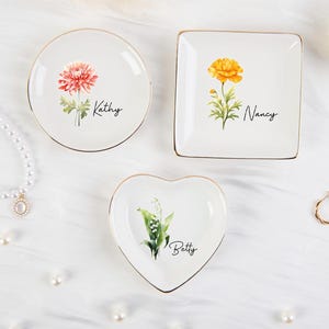 K&ouml;nnte beinhalten: Drei wei&szlig;e Schmuckschalen in runder, quadratischer und herzf&ouml;rmiger Ausf&uuml;hrung, jeweils mit floralem Design und Namen: Kathy, Nancy und Betty. Die Schalen haben goldfarbene R&auml;nder. Eine Perlenkette und ein Ring sind ebenfalls im Bild zu sehen.