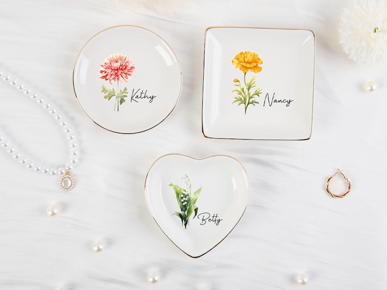 K&ouml;nnte beinhalten: Drei wei&szlig;e Schmuckschalen in runder, quadratischer und herzf&ouml;rmiger Ausf&uuml;hrung, jeweils mit floralem Design und Namen: Kathy, Nancy und Betty. Die Schalen haben goldfarbene R&auml;nder. Eine Perlenkette und ein Ring sind ebenfalls im Bild zu sehen.