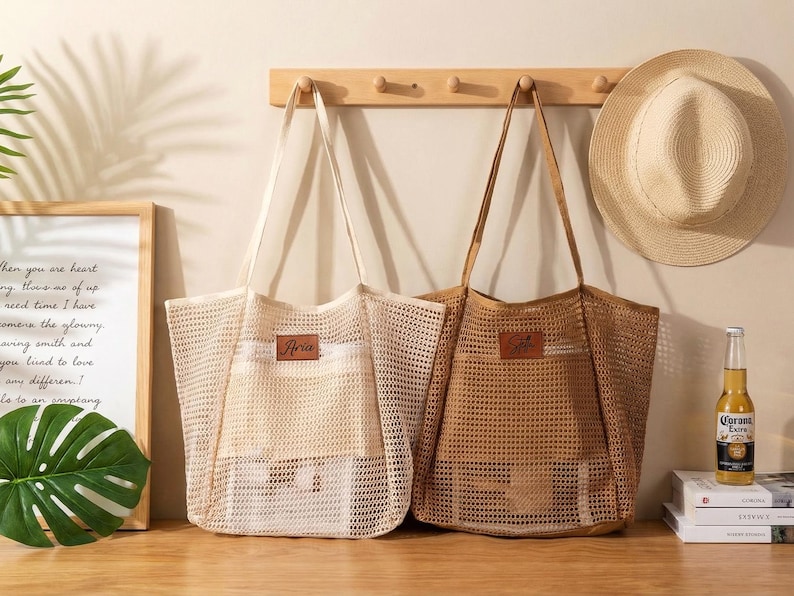 Pode incluir: Duas sacolas de rede, uma bege e outra marrom, com al&ccedil;as longas, penduradas em um cabide de madeira. Cada bolsa tem um patch de couro com o nome "Aria". Um chap&eacute;u de palha e uma garrafa de cerveja tamb&eacute;m s&atilde;o vis&iacute;veis.