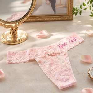K&ouml;nnte beinhalten: Ein hellrosa Spitzen-String mit einer kleinen Schleife in der Mitte und dem Schriftzug Bad Girl, der in dunklerem Rosa auf den Bund gestickt ist. Das Kleidungsst&uuml;ck besteht aus floral gemusterter Spitze mit gewellten Kanten am Bund und Beinausschnitt.