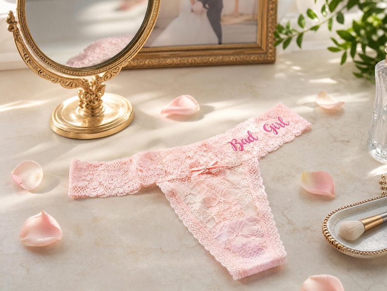 K&ouml;nnte beinhalten: Ein hellrosa Spitzen-String mit einer kleinen Schleife in der Mitte und dem Schriftzug Bad Girl, der in dunklerem Rosa auf den Bund gestickt ist. Das Kleidungsst&uuml;ck besteht aus floral gemusterter Spitze mit gewellten Kanten am Bund und Beinausschnitt.