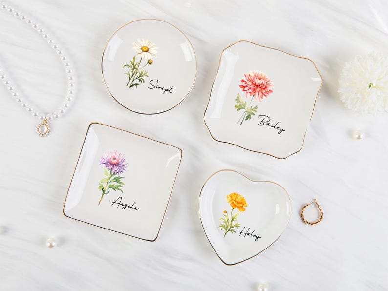 K&ouml;nnte beinhalten: Vier wei&szlig;e Keramik-Schmuckschalen mit goldenen R&auml;ndern, jede mit einem anderen Blumendesign und einem Namen: Script, Bailey, Angela und Haley. Eine Perlenkette und ein goldener Ring sind ebenfalls vorhanden.