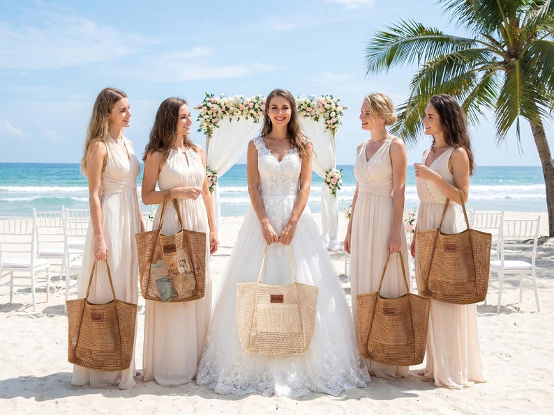 Pode incluir: Uma noiva e damas de honra numa praia, cada uma segurando uma sacola de tecido. As sacolas s&atilde;o de cor castanho natural com formato retangular e um patch de couro. As mulheres usam vestidos longos e esvoa&ccedil;antes de cor clara. Um arco de casamento est&aacute; ao fundo.