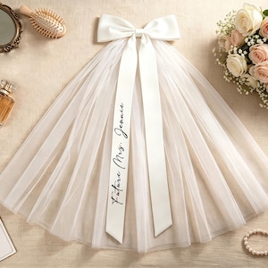 May include: A white tulle veil featuring a large satin bow at the top with two long, flowing ribbons. One ribbon is printed with the black cursive text "Future Mrs. Jennie". The veil is sheer, layered, and has a soft, gathered appearance.