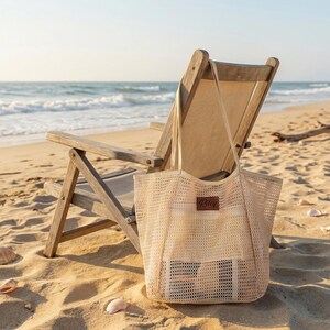 Pode incluir: Uma sacola de malha bege com um patch de couro, em uma praia arenosa ao lado de uma cadeira dobr&aacute;vel de madeira. O oceano e o c&eacute;u s&atilde;o vis&iacute;veis ao fundo, com conchas espalhadas.