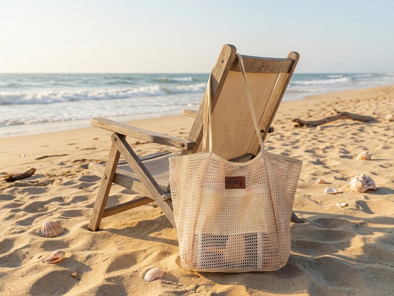 Pode incluir: Uma sacola de malha bege com um patch de couro, em uma praia arenosa ao lado de uma cadeira dobr&aacute;vel de madeira. O oceano e o c&eacute;u s&atilde;o vis&iacute;veis ao fundo, com conchas espalhadas.