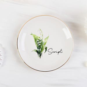 K&ouml;nnte beinhalten: Ein wei&szlig;er Keramikteller mit Goldrand, mit einer Aquarellillustration von Maigl&ouml;ckchenbl&uuml;ten und -bl&auml;ttern. Das Wort "Script" ist in schwarzer Schrift geschrieben. Eine Perlenkette und eine einzelne Perle sind ebenfalls sichtbar.