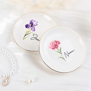 K&ouml;nnte beinhalten: Zwei wei&szlig;e Keramikschalen mit goldenen R&auml;ndern. Eine Schale zeigt eine lila Iris und den Namen "Jen", die andere eine rosa Nelke und den Namen "Naomi". Dekoratives Blumendesign.