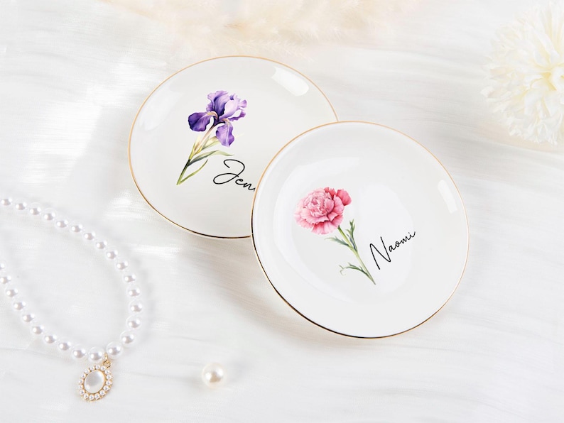 K&ouml;nnte beinhalten: Zwei wei&szlig;e Keramikschalen mit goldenen R&auml;ndern. Eine Schale zeigt eine lila Iris und den Namen "Jen", die andere eine rosa Nelke und den Namen "Naomi". Dekoratives Blumendesign.