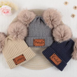 May include: Three ribbed knit beanies in beige, gray, and navy blue, each featuring two large faux fur pom-poms and a rectangular brown leather patch. The patches are engraved with the names Linsey, Pamela, and Rachel respectively.
