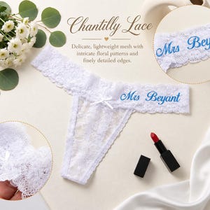 K&ouml;nnte beinhalten: Ein wei&szlig;er Spitzen-Tanga mit einer kleinen mittigen Schleife und dem in Blau gestickten Text Mrs Beyant. Das Kleidungsst&uuml;ck wird zusammen mit einem Strau&szlig; wei&szlig;er Blumen, einem roten Lippenstift und einem runden Ausschnitt gezeigt.