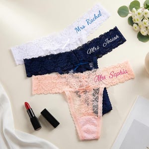 K&ouml;nnte beinhalten: Drei Spitzen-Tangas in Wei&szlig;, Marineblau und Hellrosa. Jeder hat eine kleine Schleife in der Mitte und gestickten Text: Mrs Rachel in Blau, Mrs Josiah in Wei&szlig; und Mrs Sophia in Pink. Die Kleidungsst&uuml;cke bestehen aus floraler Spitze.