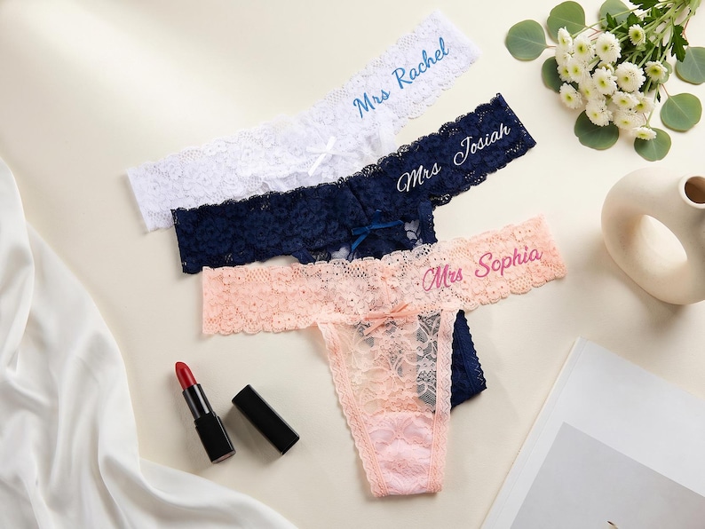 K&ouml;nnte beinhalten: Drei Spitzen-Tangas in Wei&szlig;, Marineblau und Hellrosa. Jeder hat eine kleine Schleife in der Mitte und gestickten Text: Mrs Rachel in Blau, Mrs Josiah in Wei&szlig; und Mrs Sophia in Pink. Die Kleidungsst&uuml;cke bestehen aus floraler Spitze.