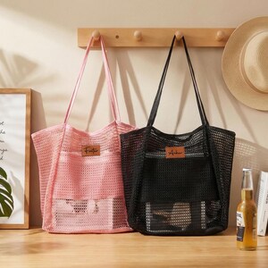 Pode incluir: Duas sacolas de rede, uma rosa e outra preta, com al&ccedil;as longas. A sacola rosa tem uma etiqueta "Foster", e a sacola preta tem uma etiqueta "Archer". Um chap&eacute;u de palha est&aacute; pendurado em um cabide de madeira acima das sacolas.