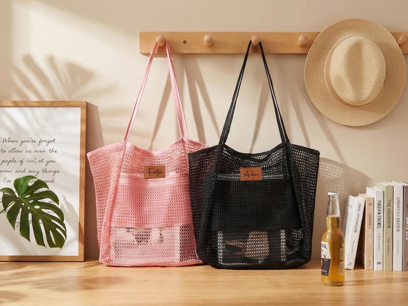 Pode incluir: Duas sacolas de rede, uma rosa e outra preta, com al&ccedil;as longas. A sacola rosa tem uma etiqueta "Foster", e a sacola preta tem uma etiqueta "Archer". Um chap&eacute;u de palha est&aacute; pendurado em um cabide de madeira acima das sacolas.