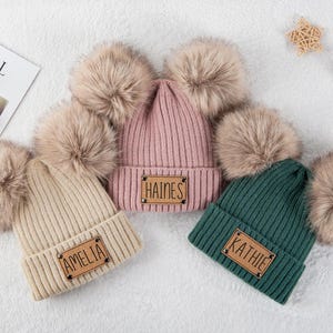 Könnte beinhalten: Drei gerippte Strickmützen mit Kunstfell-Bommeln. Eine ist beige mit "AMELIA" auf einem Lederpatch, eine ist rosa mit "HAINES" und eine ist grün mit "KATHIE". Eine sternförmige Dekoration und eine Holzrassel sind im Hintergrund.