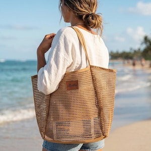 Pode incluir: Uma grande sacola tote de tecido aberto em cor bege natural. A sacola tem formato retangular com um patch de couro e al&ccedil;as longas. A sacola &eacute; carregada em uma praia com o oceano ao fundo.