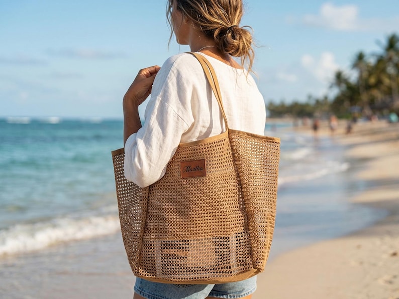 Pode incluir: Uma grande sacola tote de tecido aberto em cor bege natural. A sacola tem formato retangular com um patch de couro e al&ccedil;as longas. A sacola &eacute; carregada em uma praia com o oceano ao fundo.